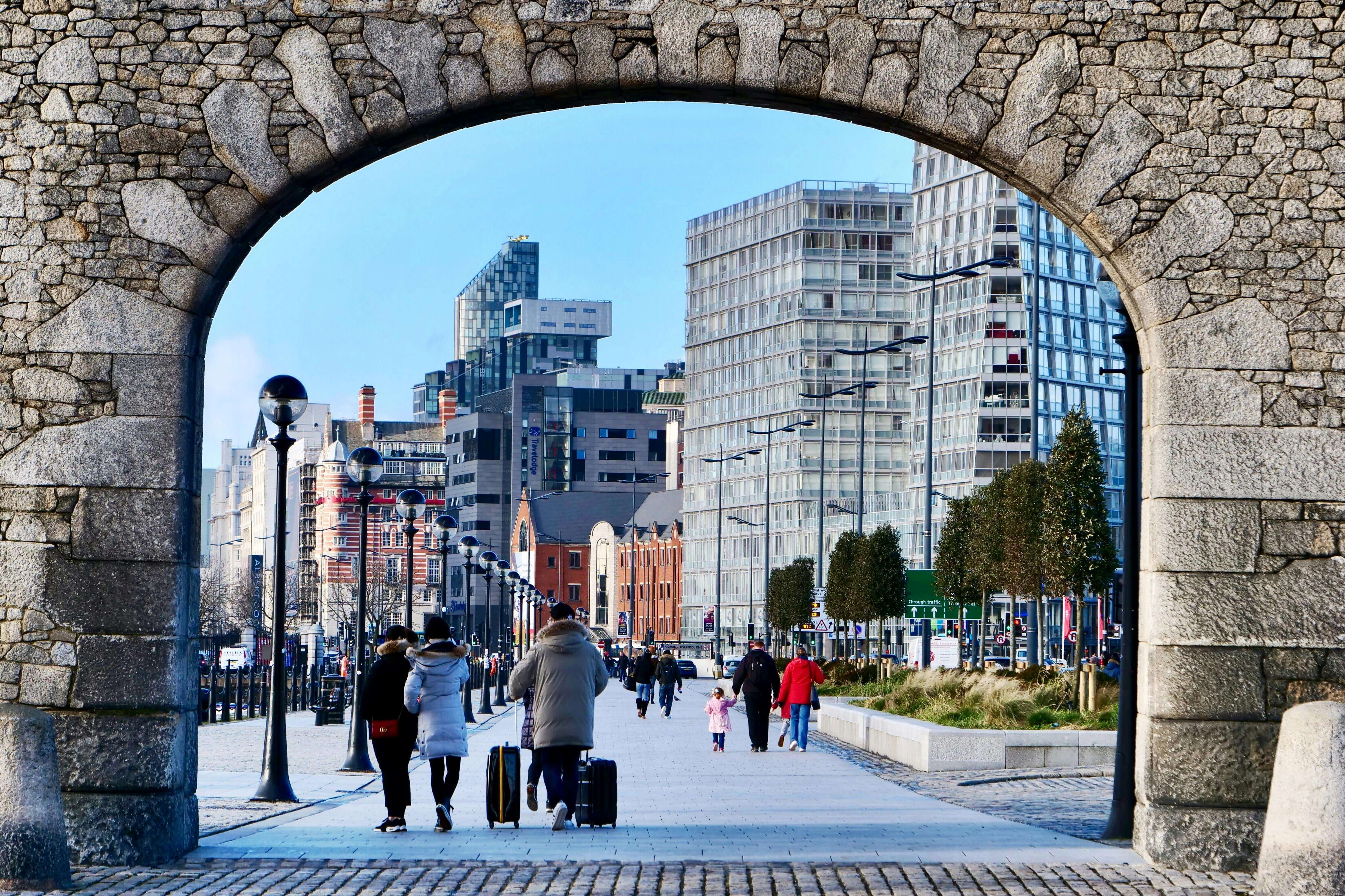 Historic stone archway framing modern Liverpool cityscape with pedestrians on waterfront promenade